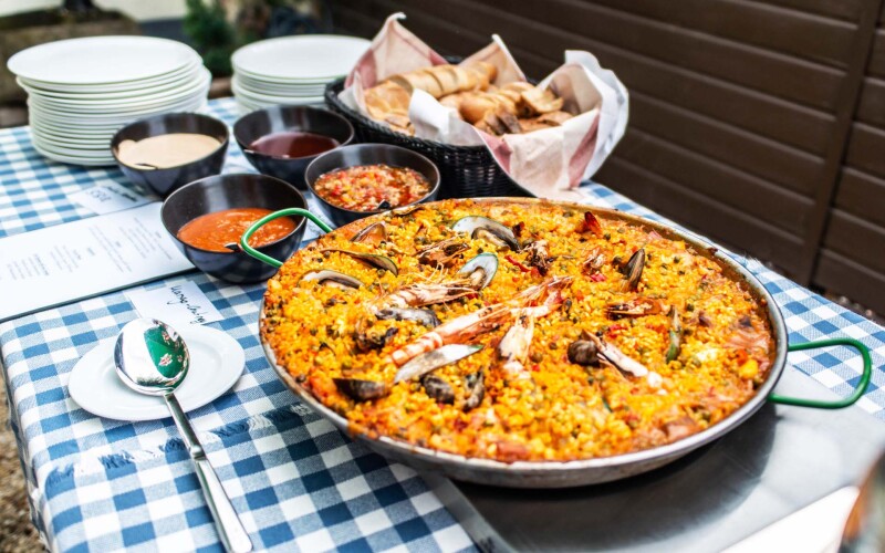 Un tavolo con sopra la paella, piatto spagnolo, con pane e salse
