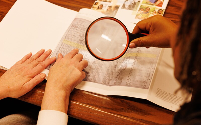 A hand points at family history records while a woman looks at the records through a magnifying glass.