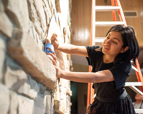 Foto van Dana Tanamachi die, op een ladder, tape aan de muur plakt.