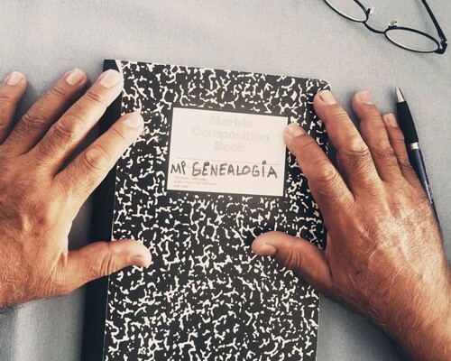 Hai bàn tay cầm một cuốn sổ có ghi chữ “mi genealogia” trên đó.