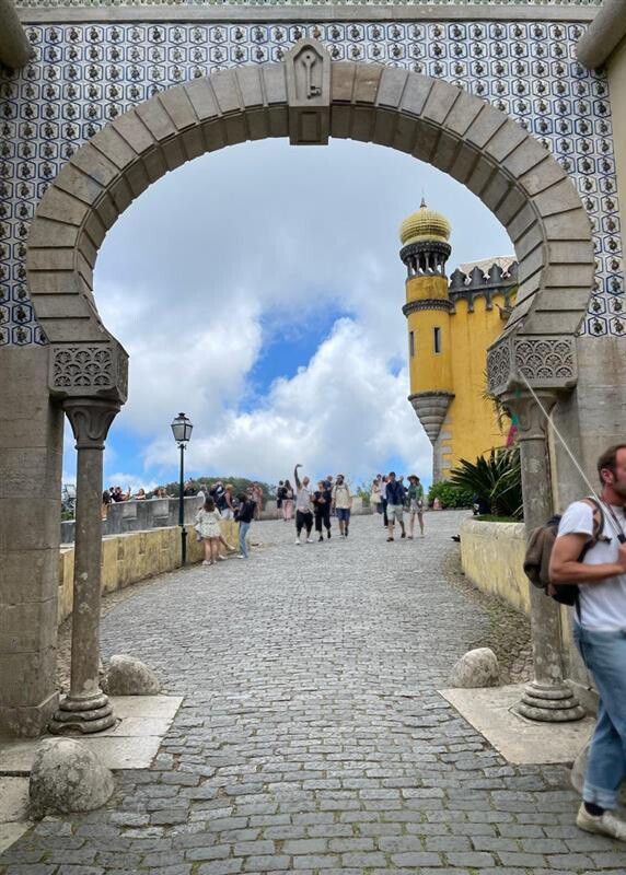 turisták sétálnak egy lisszaboni utcán, egy gyönyörű boltív alatt elhaladva