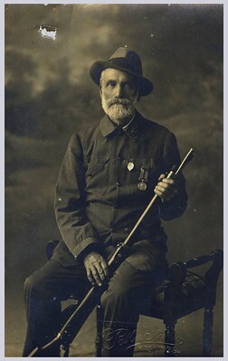 William Heslop, New Zealand Wars Veteran, posed in uniform, sitting and holding military rifle.