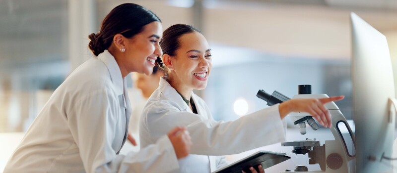 Two female scientists in lab coats smiling while looking at a computer screen.