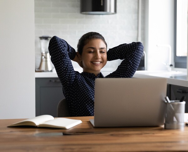 Eine junge Frau sitzt entspannt und glücklich am Computer, auf dem Schreibtisch liegt ein Buch