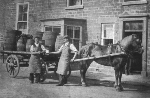 A black-and-white photograph of two draymen standing next to a dray attached to a horse carrying barrels of goods.