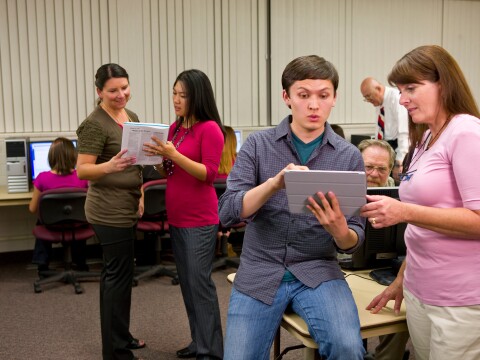 A woman assists a young man doing family history on his tablet while others sit at desks and work on computers.