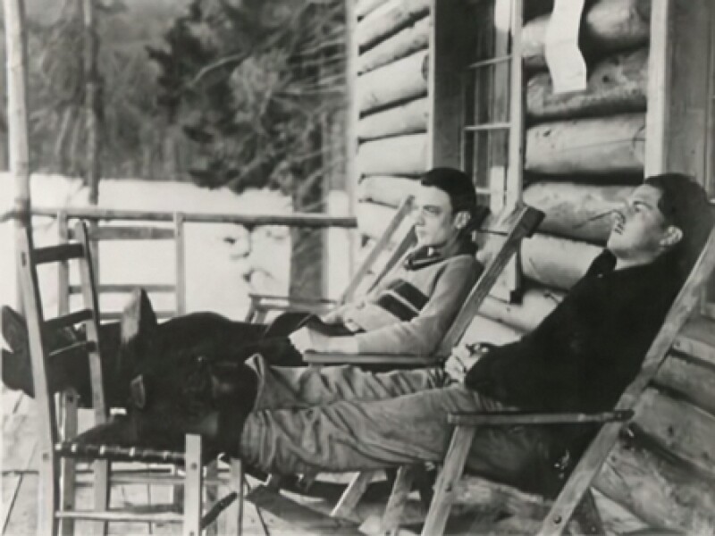 1931 photo from Canada of two men sitting in reclined wooden chairs with their feet up, in front of a log cabin home.